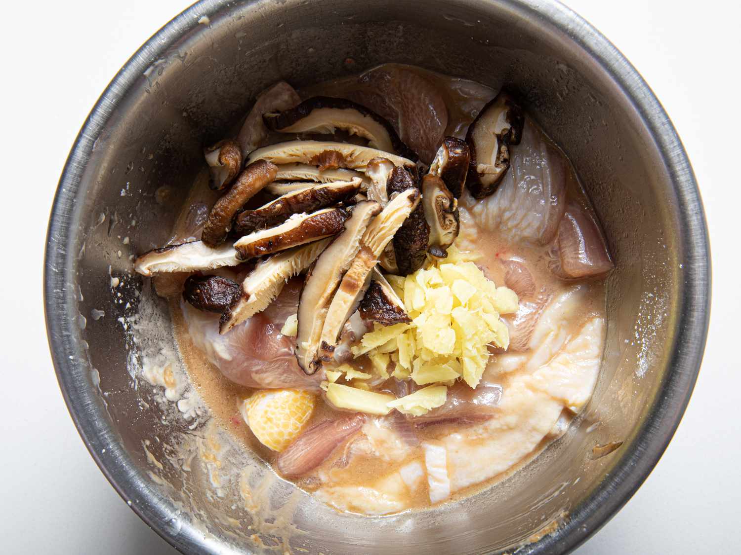 Shiitake mushrooms and ginger on top of raw chicken in a stainless steel bowl.