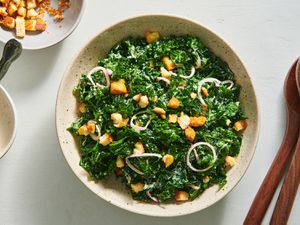 Kale caesar salad in a speckled ceramic bowl. On the right periphery are a pair of wooden serving spoons, and on the left periphery is a small bowl holding croutons.