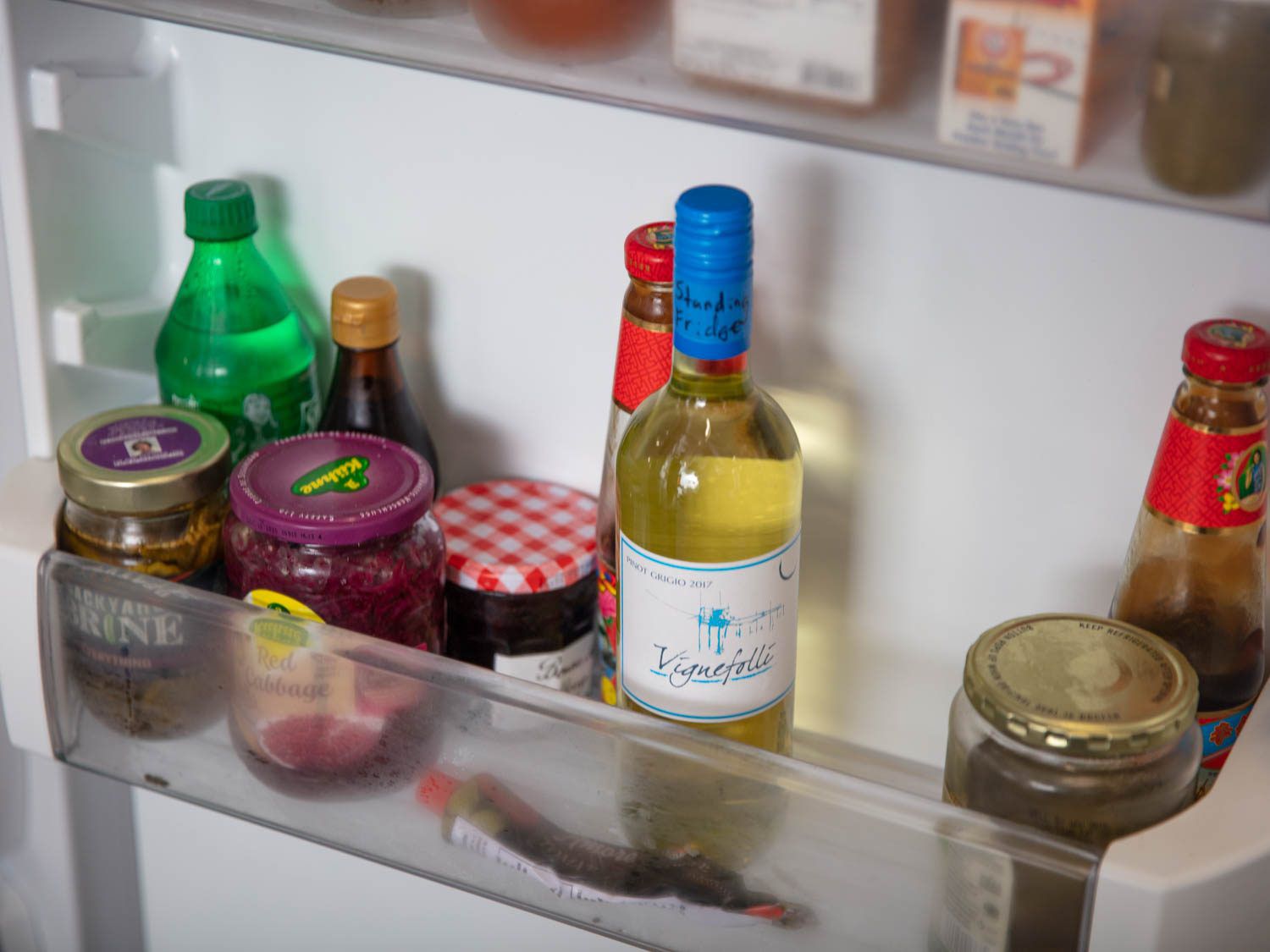 A bottle of white wine upright in the door of a refrigerator, next to a jar of jam and other condiments.