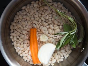 A pot filled with dry beans, a carrot, a garlic clove, onion, and a sprig of fresh sage.