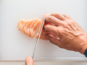 Two pairs of hands, one holding a knife and the other securing the chicken breast on a cutting board, with a chicken breast being thinly sliced.