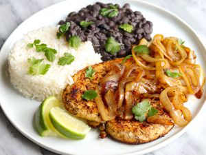 A plate of Cuban-style pollo a la plancha topped with caramelied onions, and served with white rice, black beans, lime wedges, and cilantro.