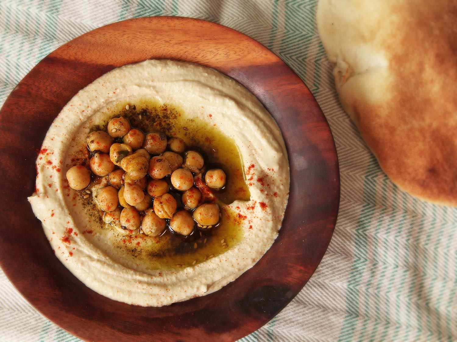 Overhead photograph of smooth Israeli-style hummus topped with chickpeas, olive oil, za'atar, and paprika, with pita bread in the background.