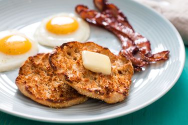 Split homemade English muffin topped with butter on a blue plate next to two sunny-side-up eggs and two slices of bacon.