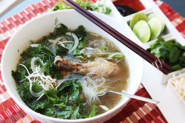 White bowl of pho ga on red placemat with chopsticks and a spoon on the bowl and garnishes on the side.