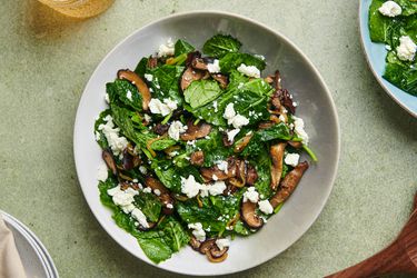 Warm kale and caramelized mushroom salad in a white ceramic bowl on a green surface. There are a pair of wooden spoons at the bottom right corner, a glass of golden liquid in the top left corner, a stack of plates covered with a napkin in the bottom left corner, and another plate of salad in the top right corner.