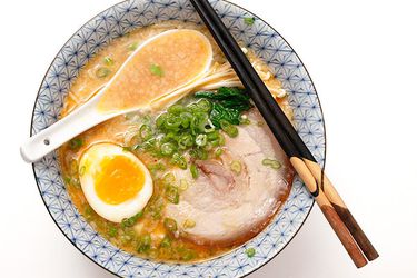 A bowl of tonkatsu ramen with half a soft-boiled eggs, green onions, and a slice of pork on top. A Chinese soup spoon and a pair of chopsticks are sitting on the rim of the bowl.