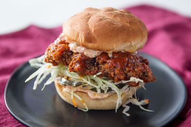 A kimchi-brined fried chicken sandwich with cabbage, pickles, and sauce on a dark plate.