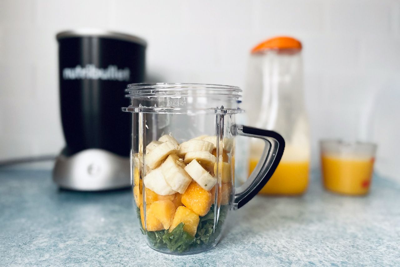 香蕉、冷冻芒果和羽衣甘蓝放在一个搅拌器罐中，搅拌机和橙汁作为背景。