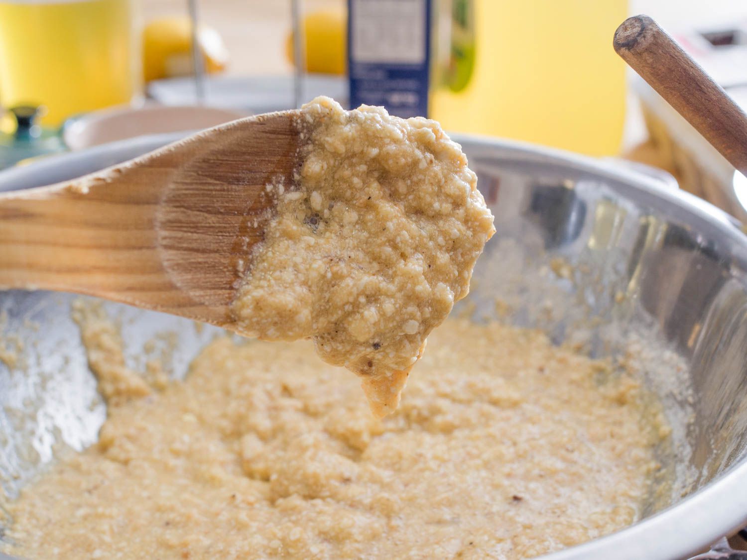 A wooden spoon held up and tilted to the side over the mixing bowl. Runny, just-mixed matzo ball batter is dripping back into the bowl.