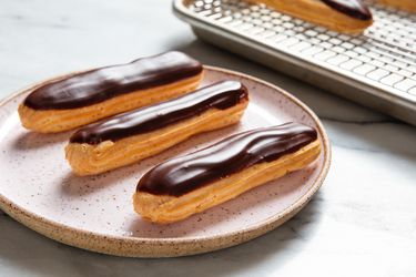 小粉红斑点炻器平台e with three classic éclairs on it, each topped with chocolate icing. A bakers rack with more éclairs is in the background.