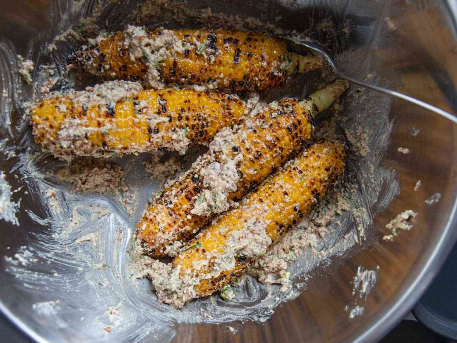 Ears of grilled corn being tossed with mayonnaise, crema, Cotija cheese, scallions, and XO sauce in a large mixing bowl.