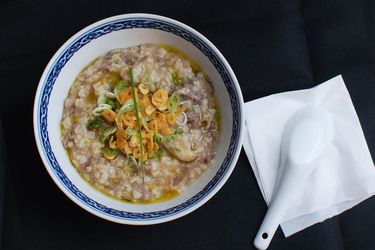 Overhead view of a bowl of brown rice congee with beef, shiitake, and garlic chips, served on a black table with a soup spoon.