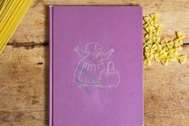 A copy of the book Strega Nona by Tomie dePaola, on a wood tabletop with dried pasta. .