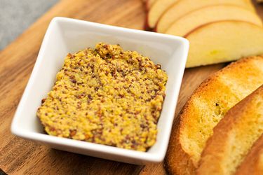 Closeup of a ramekin of whole grain Dijon mustard, set next to toasted baguette and apple slices.