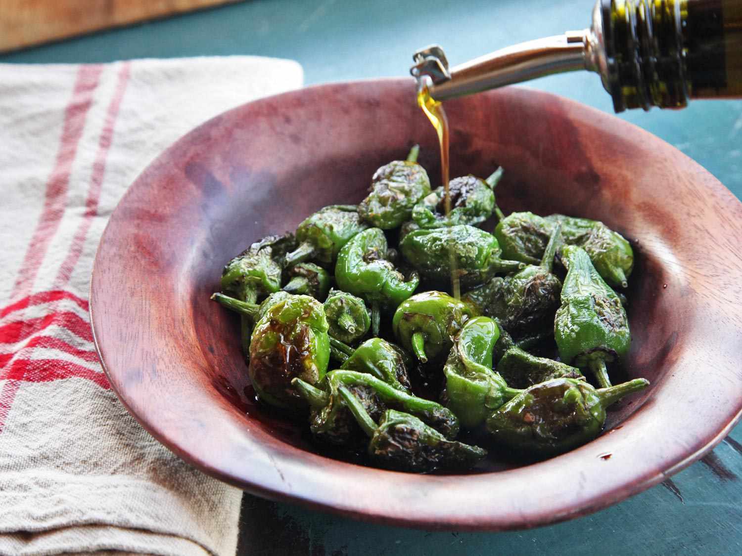 Olive oil being poured over cooked Padrón peppers in a bowl,