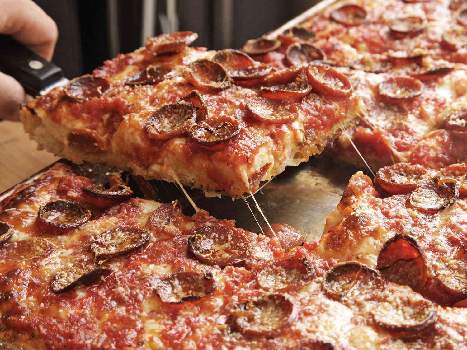 Removing a slice of pepperoni-topped Sicilian-style pizza from a sheet pan.