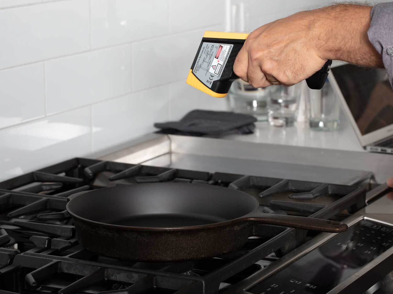 The reviewer uses an infrared thermometer to measure heat on the surface of a cast iron skillet over a moderate flame.