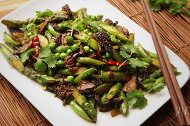 Stir-fried spring vegetables with black olives and Sichuan peppercorns, served on a square platter.