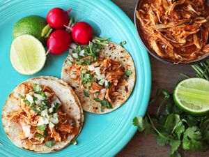 Two street tacos topped with chicken tinga, diced onion, and cilantro. There are radishes and cut limes on the plate and a dish of chicken tinga nearby.