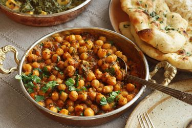 Channa masala in a metal serving dish next to a plate of naan bread and a dish of palak paneer.