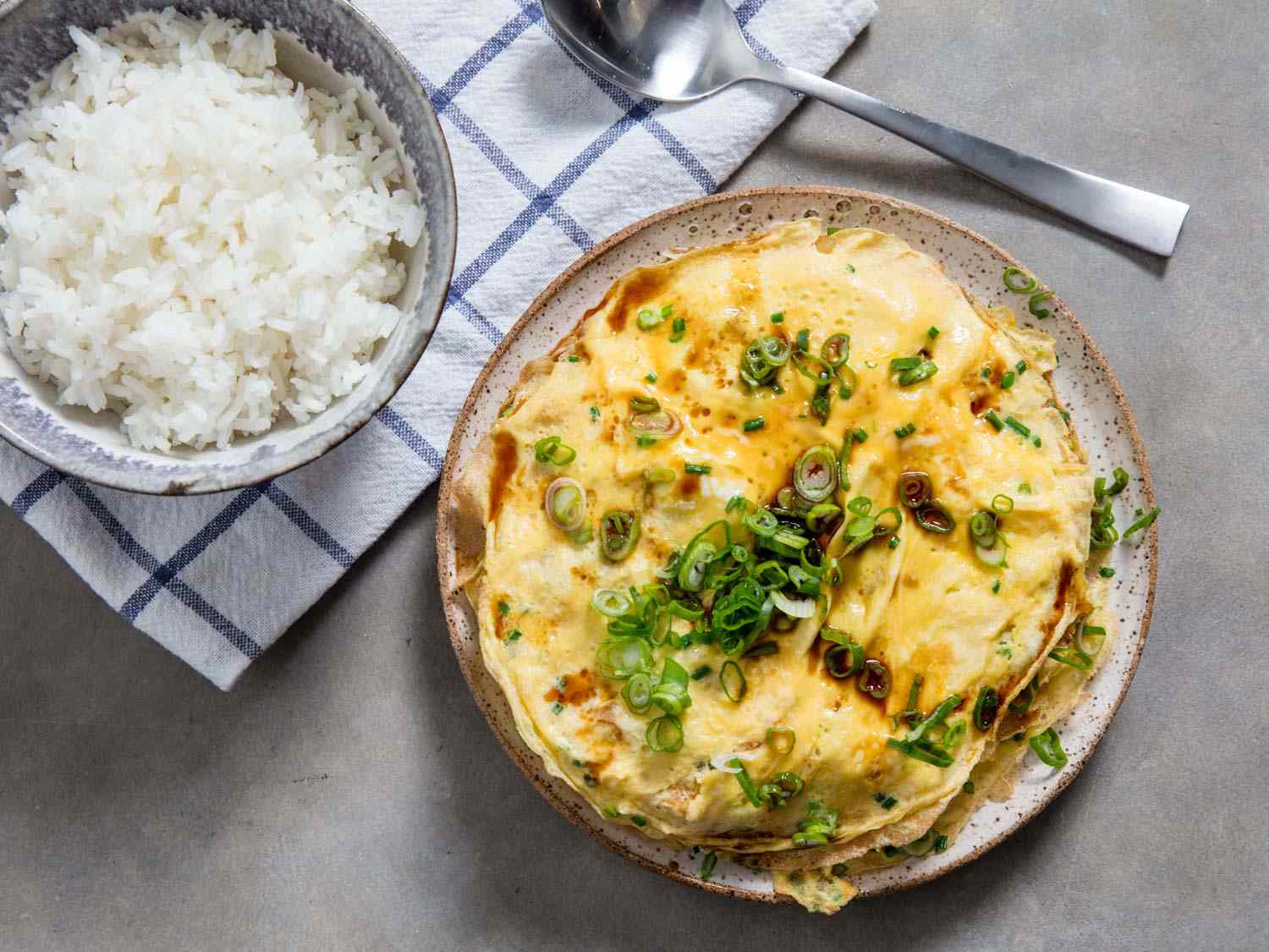 An overhead view of a layered omelette topped with scallions next to a bowl of rice.