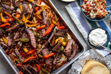 A sheet pan loaded with skirt steak strips, bell peppers, and onions, all after being cooked until charred, ready to be loaded into tortillas for fajitas