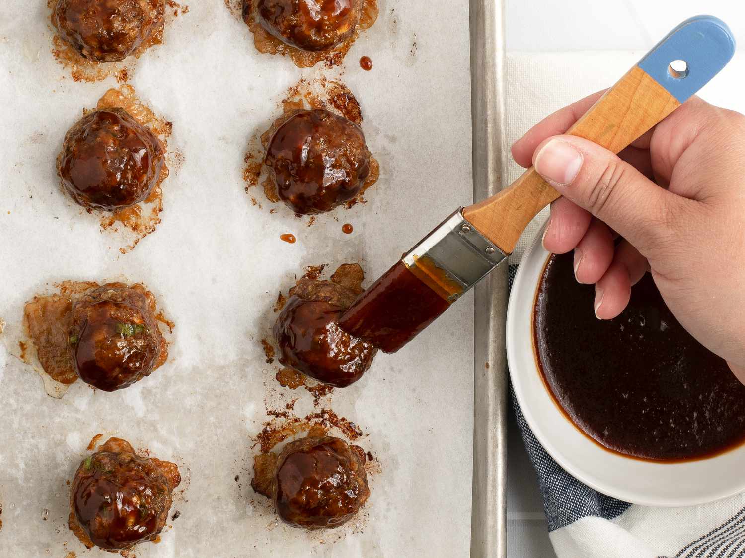 The cooked meatballs, on parchment paper inside of a baking sheet, being brushed with the cooked hoisin sauce.