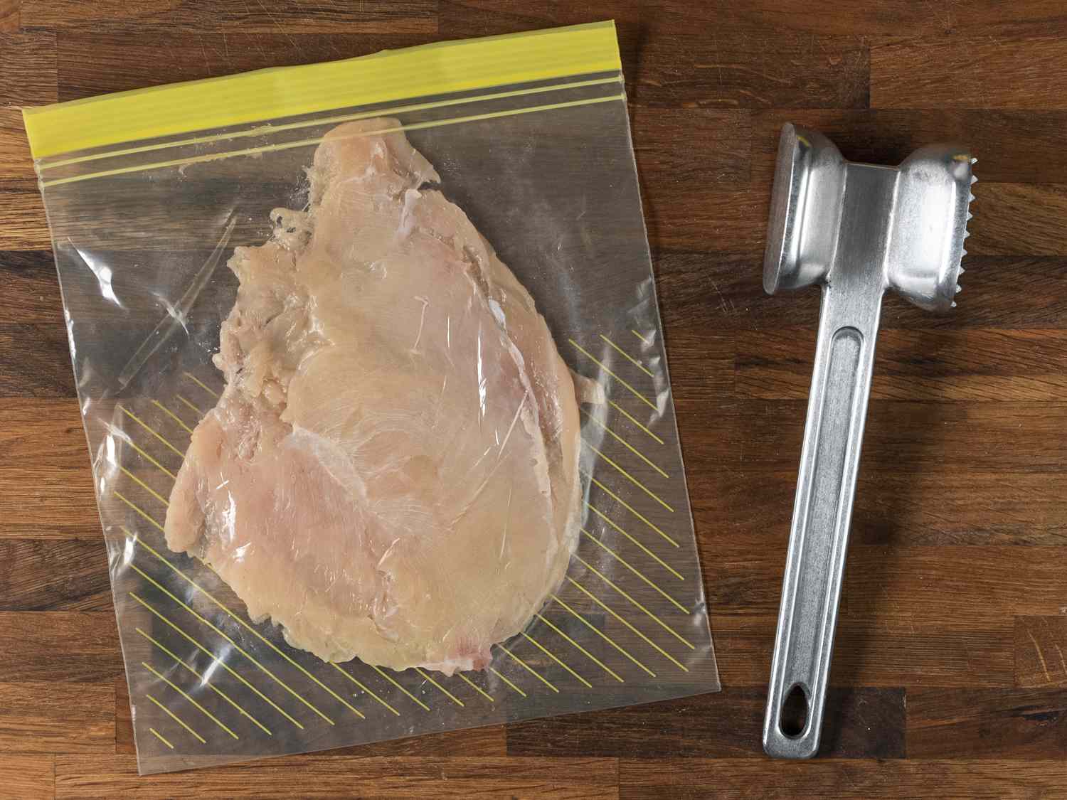 鸡胸肉放在一个密封的拉链锁袋里，捣碎成四分之一英寸的厚度，袋子旁边有一个金属肉捣碎器。袋子和锤子都放在木制的背景上。