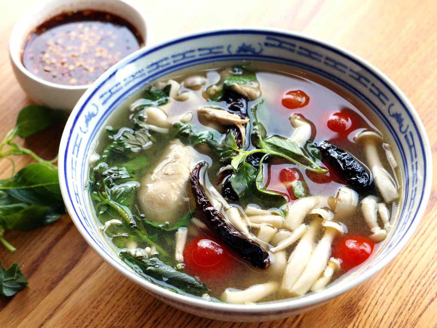 一碗蓝白相间的khao soi(鸡汤里的宽米粉、西红柿、蘑菇和绿色蔬菜)，旁边放着一小碗辣椒酱