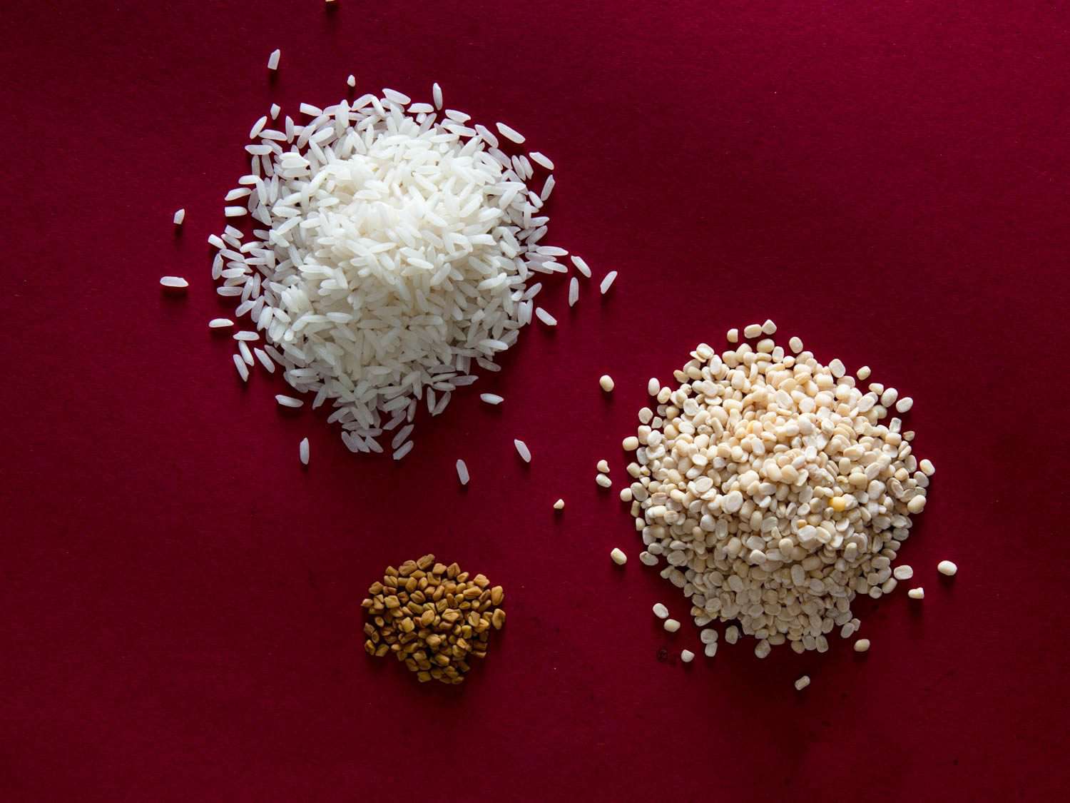 Piles of long-grain rice, urad dal, and chana dal against a dark red background.