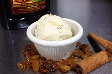 A scoop of rum raisin ice cream served in a ramekin. A bottle of dark rum, golden raisins, and cinnamon sticks are nearby.