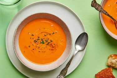 A bowl of Andalusian gazpacho in a ceramic bowl set on a plate. On the right hand periphery are another bowl of soup and a couple of pieces of torn bread.