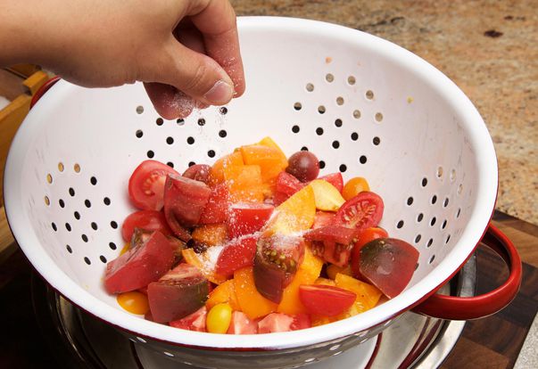 A hand sprinkling salt over chunks of yellow and red ripe tomato, in a colander placed over a bowl