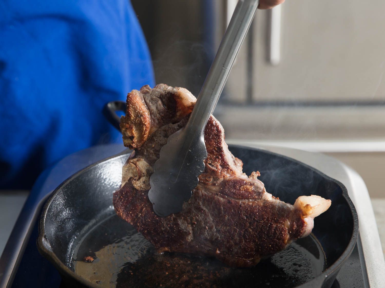 Tongs turning a steak that's cooking in a cast iron skillet