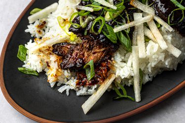 A close-up of slow-cooked Korean-inspired short ribs with green onion and pear, plated with white rice on a black oval ceramic dish.