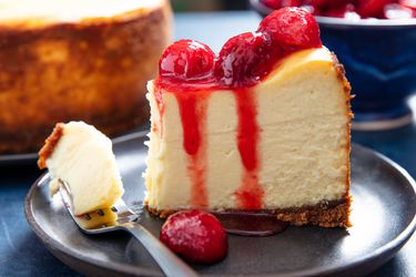一块史诗般的纽约芝士蛋糕放在盘子里，上面浇上樱桃酱汁。＂width=