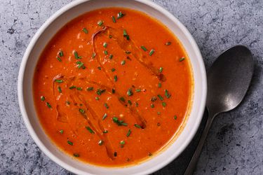 一碗奶油素食番茄汤在石头的背景上，勺子在碗的右边。汤的表面淋上橄榄油，撒上切碎的香草。