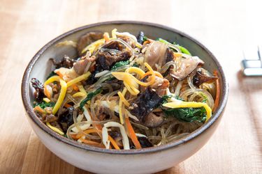 A bowl of jap chae showing colorful egg strips, spinach, carrot, and more intertwined in the noodles.