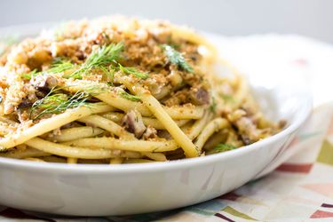 A shallow white bowl filled with a mound of pasta con sarde topped with bread crumbs.