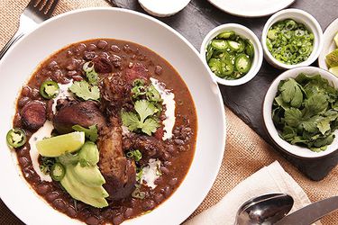 Bowl of black bean soup with large pieces of meat, avocados, and jalapenos on top.