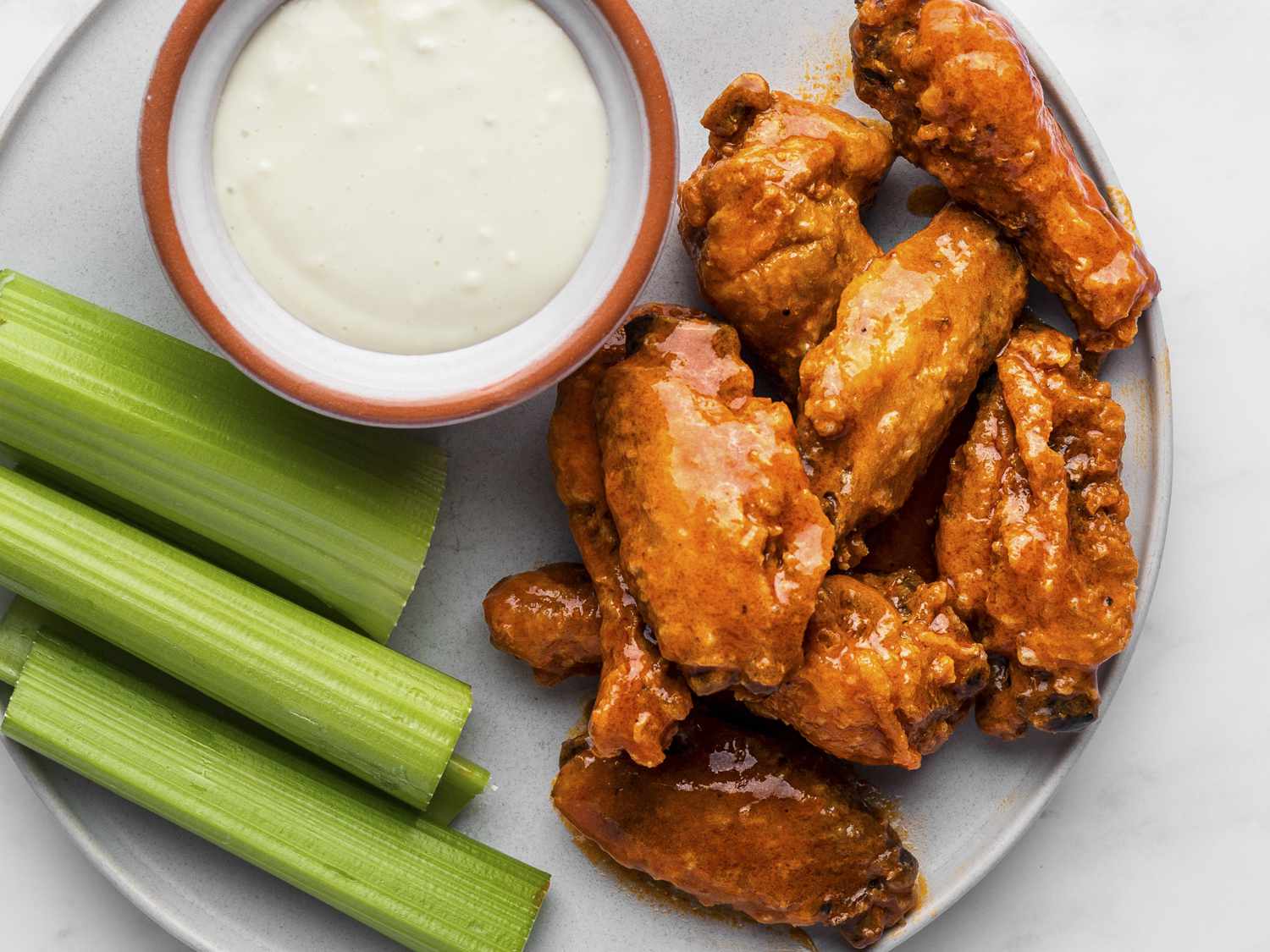 Crispy, sauce-covered fried buffalo wings on a white ceramic plate with sliced celery sticks and a small bowl of bleu cheese dressing, on a white stone background.