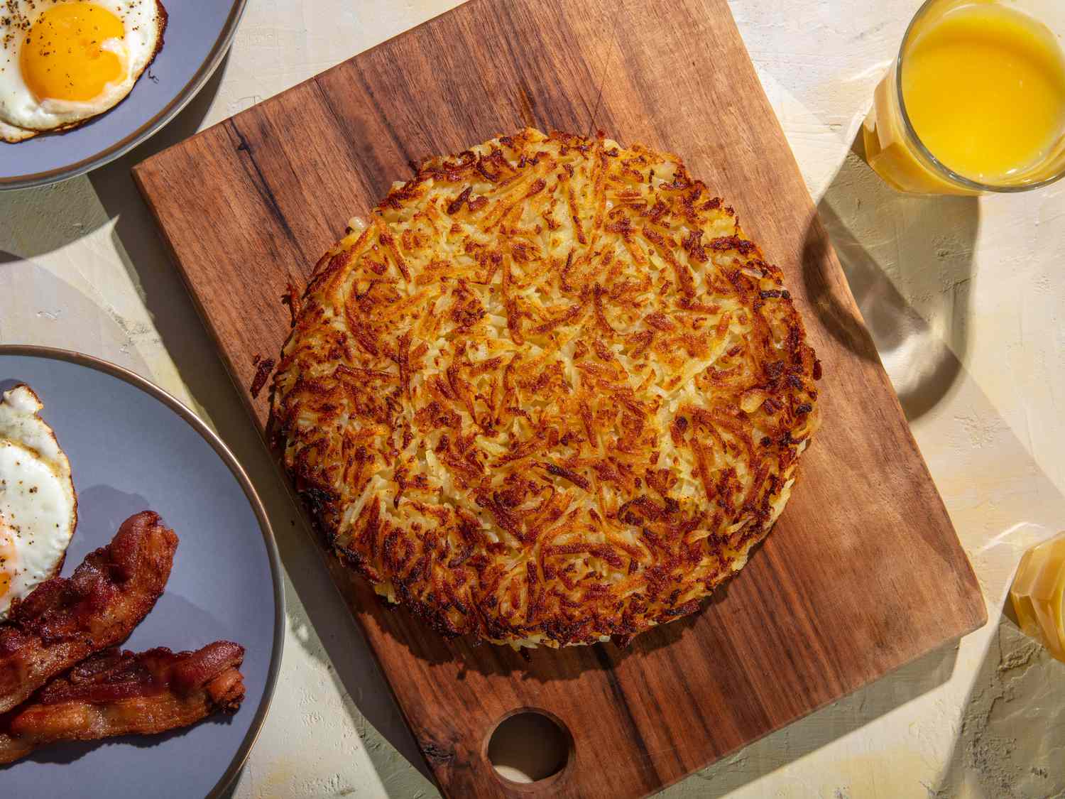 A deeply browned rösti sits on a wooden cutting board, with plates of fried eggs and bacon, and glasses of orange juice around it on the table.