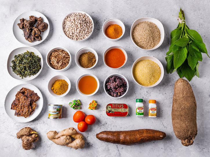 Overhead view of all the essential ingredients needed in a pantry to cook a range of Nigerian dishes. Ingredients range from spices to roots to tinned food and fresh spices, organized in a neat grid