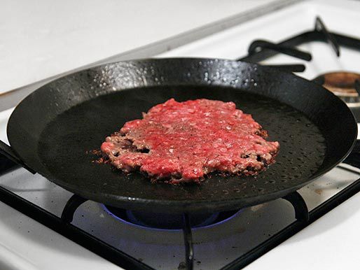 完全粉碎的汉堡肉饼放在冒烟的热碳钢煎锅上。＂width=